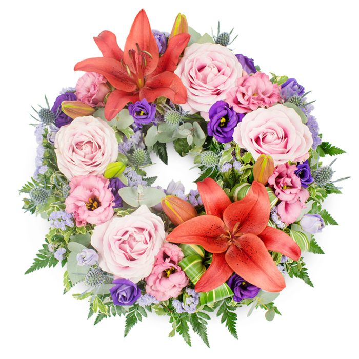 A beautifully composed funeral wreath is displayed flat against a clean white background, photographed from directly above so that its circular form is perfectly framed. The arrangement feels both abundant and carefully controlled, each element thoughtfully placed to honour a loved one with dignity. Two striking Asiatic lilies in a rich coral-orange tone form the main focal points; one is situated near the upper curve of the wreath and the other nearer the lower right side, their broad, star-shaped petals splaying outward and their stamens creating a touch of intricate detail at the centre. Between these lilies, numerous roses in soft blush and lavender-pink tones are nestled closely, their fully opened, spiralling petals giving the wreath a romantic and soothing presence. Lisianthus (or eustoma) flowers are woven throughout in complementary shades, from pale, almost pastel pink to deep, velvety purple, their ruffled, cup-like blooms adding layers of texture that resemble gently folded fabric. Slim, sculptural eryngium thistles in a blue-grey hue punctuate the design, bringing an element of contrast that is both natural and slightly architectural. Wispy sprays of tiny lavender-blue filler flowers, such as statice or limonium, drift between the larger blooms, creating a soft, misty backdrop. All of this is supported by a lush base of greenery, with silvery eucalyptus leaves, feathery ferns, and other textured greens overlapping into a dense, verdant ring. A few unopened lily buds peek from the foliage, symbolising remembrance and life continuing beyond farewell. Near one lily, neatly folded bright green leaves reveal skilful craftsmanship you might expect from a Highgate florist preparing flowers for a memorial near Highgate Cemetery or overlooking the views by Highgate village. The overall mood is peaceful, respectful, and deeply compassionate.