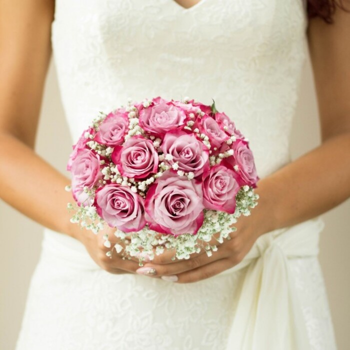 Drawing the viewer into a moment of joyful anticipation, this detailed shot captures a bride from the chest down, cherishing a bouquet that radiates both modern sophistication and Highgate tradition. A tightly gathered cluster of two-tone pink roses forms the core, each flower unfurling from a soft, gentle blush at its centre to vivid outer petals of magenta and fuchsia-creating an effect of luminous depth beloved by local florists. Interspersed throughout, delicate clouds of white baby's breath evoke the soft mist that often drifts across Highgate Wood on early mornings, lending an air of enchantment to the arrangement. Hints of green from neatly trimmed stems peek through, while the bride's hands, manicured in shimmering nude, frame the scene with understated grace. The surrounding white wedding gown, its bodice adorned with intricate lace in stylized floral patterns, is finished with a gently draped sash, echoing the elegant style often seen in Highgate's timeless wedding venues. The ensemble stands as an emblem of meticulous craftsmanship and heartfelt romance-speaking to any bride dreaming of a bouquet rooted in local beauty and tradition.