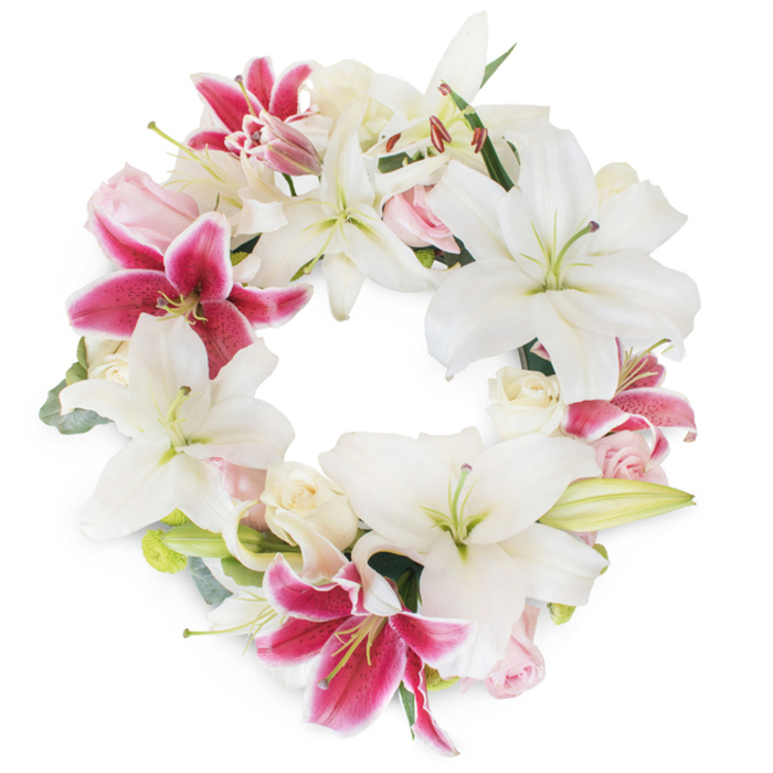 A meticulously arranged circular wreath, glimmering with light on a clear white background, celebrates remembrance and artistry inspired by Highgate's green spaces and historic charm. The focal point is a lavish gathering of large, white Oriental lilies-some open wide to reveal vivid green pistils and golden stamens-each petal edged in soft shadow and pure light. These are contrasted by fuchsia-pink Stargazer lilies whose deep magenta centres and gentle speckles bring vibrant drama to the design, evoking Highgate's seasonal gardens in bloom. Clusters of delicate roses-cream-white and gentle blush-are tucked thoughtfully between the larger lilies, with some petals still curled in promise, others unfurled in quiet tribute. Interspersed tendrils of eucalyptus-like foliage and subtle green sprigs echo the area's woodland fringes, adding structure, depth, and a soothing, fresh presence. The circle is full, seamless, and balanced, communicating eternity and the enduring power of love. Bathed in natural light, each flower appears luminous and tangible, casting luminous shadows against the clean white field. This wreath makes an ideal centrepiece for a funeral or memorial in Highgate, expressing sympathy and heartfelt remembrance with a blend of vibrant colour and soft serenity-a signature of thoughtful local floristry in this cherished North London enclave.