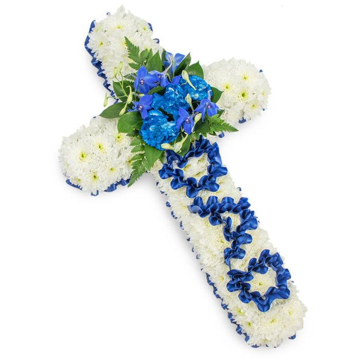 The image shows a lovingly crafted funeral cross, photographed on a seamless white background so that the focus is entirely on the flowers and detail of the design. The cross itself is generously filled with white spray chrysanthemums, each small bloom fresh and open, creating a continuous layer of petals with a soft, almost velvety appearance. The flowers are arranged so densely that the structure beneath is invisible, giving the impression of a solid cushion of white. Framing this serene base is a border of royal blue satin ribbon that runs smoothly along the outside of the cross. The ribbon is rich in colour, a deep cobalt blue that contrasts strongly with the white, while its satin finish adds a gentle lustre that hints at candlelight or the subdued glow of a chapel interior in Highgate. At the centre where the arms meet, a lush grouping of royal blue flowers forms a vivid heart to the design. Ruffled blue carnations sit alongside more delicate, upright blooms reminiscent of delphiniums or orchids, their shapes and tones adding texture and height. These are nestled into dark green leaves and fern fronds, which reach outwards and soften the intersection. Along the length of the vertical arm, the word DAD is spelled with loops of the same blue satin ribbon, each letter carefully constructed and placed to stand out against the white chrysanthemum background without disturbing its smoothness. The overall mood is one of peaceful remembrance and devoted respect, well suited for a funeral or graveside service around Highgate, perhaps near Highgate Cemetery or a local parish church, where families seek a thoughtful, personalised floral tribute that honours a father's life and love.