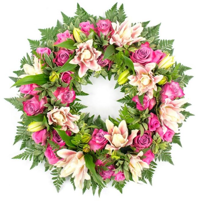 Photographed head-on against a bright white background, this circular sympathy wreath reveals a meticulously crafted blend of flowers and foliage, every inch of the ring carefully filled. The arrangement centres on a profusion of roses in rich, jewel-like shades of fuchsia and magenta softened by paler pink tones. Their petals are densely layered, some blooms still partially closed with a sense of youthful energy, others fully open and relaxed, showing the full spiral of the rose's heart. Woven through the roses are lush double lilies in creamy white and soft blush pink, with hints of peach on their slightly frilled petals. Several of the lilies are wide open, their centres clearly visible, while others are contained in lime-green buds, offering a suggestion of future bloom even in an arrangement created for farewell. The greenery plays an equal role in the composition: feathery fern fronds extend along the outer edge of the wreath, creating a soft, textured outline, while glossy, structured leaves in various shades of green fill spaces between flowers, adding depth and supporting the circular shape. Together, the colours move from cool, bright greens to warm, comforting pinks and creams, creating a visual rhythm that feels both uplifting and serene. This type of wreath feels thoughtfully suited to a Highgate service, envisioned perhaps resting at a graveside among old trees or inside a chapel near the quiet paths around Highgate Wood. The pure white background in the image accentuates the wreath's fullness and craftsmanship, underscoring a message of respect, love, and lasting remembrance.