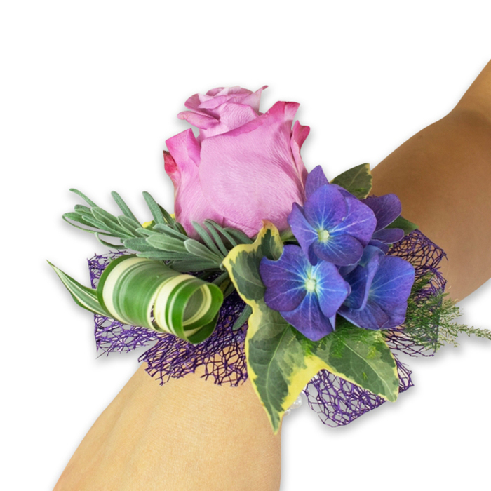Presented diagonally across a light-toned wrist against a clean white backdrop, this corsage is a study in refined contrasts: a large, partially open pink rose forms the romantic centrepiece, its petals graduating from a pale blush to a richer fuchsia at the ruffled edges, while three dense clusters of indigo-purple hydrangea flank it, each tiny floret marked by a luminous blue-white eye and a velvety surface that adds weight and visual rhythm. Strategically placed greenery creates depth-a fan of slender, silvery-green stems reminiscent of lavender or eucalyptus provides a soft, aromatic veil behind the blooms, while glossy, variegated ivy leaves with dark green centres and creamy margins lend a classic touch. A modern flourish appears as a broad, white-and-green striped leaf, meticulously curled into a spiral to introduce sculptural movement. All of this rests atop a dark purple, open-weave mesh that both anchors the composition and amplifies the purple notes of the hydrangeas, offering subtle texture against fabrics and catching light like evening tulle. As a Highgate florist who prepares many corsages for Waterlow Park weddings and graduations at nearby colleges, I craft this piece to balance durability with delicate beauty-securely wired and cushioned so the wearer can dance or greet guests without worrying about petals. The scent is faintly floral and uplifting, the hand-feel a contrast of silky rose petals, plush hydrangea, and crisp ivy, and the overall impression is celebratory and elegant. Flowers Highgate often pairs this design with simple wrist ribbons or a discreet fastening to suit formal gowns or smart tailoring, producing a memorable accent for important local occasions.