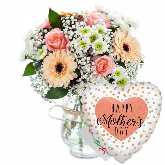 A lovingly composed Mother's Day gift arrangement is pictured against a bright, seamless white background, much like the product displays seen on Flowers Highgate's website, yet it retains a personal, handcrafted character. On the left, a full, rounded bouquet brims from a clear glass jar-style vase. The vase's neck is wrapped with a slim band of rustic twine, tied in a modest bow that suggests the touch of an experienced local florist. The bouquet showcases a palette of gentle pastels: several peach and light pink roses open to reveal their classic spiralling centres, the petals soft and velvety, catching light on their edges. Two or more large peach-toned gerbera daisies with pronounced dark brown centres stand proudly among the roses, adding a confident, upbeat note. Between these focal blooms, clusters of tiny white gypsophila weave through like delicate lace, and smaller white chrysanthemums with light green centres fill out the shape, bringing a fresh yet subtle contrast. Deep green foliage supports and encircles the flowers, adding volume and evoking nearby Highgate gardens in late spring. The stems can be clearly seen through the transparent glass, emphasising the natural, freshly cut quality of the bouquet. Nestled just behind the flowers and partially obscured is a small white greeting card, its colourful lettering slightly blurred but unmistakably meant for Mother's Day. On the lower right, a heart-shaped foil balloon overlaps the vase. It is crisp white with scattered metallic gold and silver dots, reminiscent of celebratory confetti. In the middle of the balloon, a solid peach heart frames the message HAPPY Mother's DAY in black fonts that combine bold clarity with graceful script. The composition suggests a thoughtfully chosen Mother's Day surprise, ideal for delivery to families across Highgate and surrounding areas.