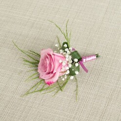 A delicately crafted pink rose boutonniere lies on a woven beige linen surface, the fabric's subtle texture forming a calm, neutral stage that lets the flowers take centre stage. In the middle, a single rose unfolds in a graceful spiral, its inner petals a soft blush that gradually deepens to richer pink tones towards the edges, creating a gentle ombré effect that speaks of careful growing and perfect timing. Around the base of this rose, sprigs of white baby's breath cluster like tiny stars, their miniature blooms adding lightness and a traditional romantic touch often favoured for weddings and proms across Highgate and nearby Muswell Hill. Behind the rose, fine strands of emerald-green, grass-like foliage sweep outward, giving the boutonniere a light, airy silhouette and suggesting the natural movement of a garden breeze in Waterlow Park. A single, broader leaf in a deeper green wraps neatly around the lower portion, folded with precision to form a smooth, tailored base that mirrors the clean lines of a formal jacket. The stems are bound together with a lustrous satin ribbon in complementary shades of lavender-pink and rich magenta, the two-toned wrapping catching the soft light and adding a polished finish, as if ready to be handed over just before a ceremony at a local Highgate church. The lighting is even and gentle, casting fine shadows that emphasise the rose's velvety texture and the baby's breath's delicate structure. This image captures the quiet elegance, tenderness, and workmanship behind a classic lapel flower designed for a day full of love and celebration.