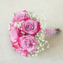 A luxurious bouquet crafted in the heart of Highgate, this wedding arrangement tells the story of refined local taste and romantic tradition. The focal roses, each unfurling velvet petals that shift from a calm lavender core to dynamic magenta edges, create depth and movement as if bathed in soft natural light common to North London's leafy mornings. Delicate sprays of white baby's breath-gypsophila-nestle in between, adding contrast and a touch of airiness, much like the gentle atmosphere found along Highgate's historic lanes. The stems are carefully gathered and wrapped snugly in a sumptuously smooth plum-coloured satin ribbon, highlighted by spirals of pristine pearls-a detail reflecting both contemporary luxury and subtle nods to Highgate's storied past. The ends of the stems remain exposed beneath the decoration, bringing a fresh, authentic feel N6 residents appreciate in locally sourced flowers. Laid atop a light beige, textured linen that enhances its gentle colour palette, the bouquet radiates quiet sophistication-ideal for brides seeking both modern style and classic grace at local venues near Waterlow Park or the Village Green. This arrangement is suited for the most significant of days, capturing that much-loved blend of celebratory elegance and understated North London charm.
