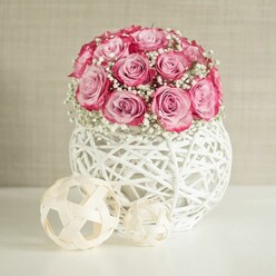 An elegant floral composition captures a rounded bouquet of pink roses and white baby's breath, styled with the quiet refinement you might expect from a Highgate village home. At the centre of the image, around fifteen roses are massed tightly together into a neat, almost spherical shape. Each rose is fully open, their petals layered and velvety, fading from a pale blush in the heart of each bloom to a more saturated, almost raspberry pink along the edges, giving a rich two-toned look. Threaded between these roses, delicate sprigs of gypsophila scatter clusters of tiny white flowers, like soft foam resting on a sea of pink, adding both volume and a sense of lightness. The bouquet emerges from a distinctive spherical vase, woven from white-painted fibres in an airy, criss-cross pattern that resembles a modern wicker lantern. Light filters through the gaps in the vase, casting subtle shadows on the pristine white surface beneath, which could be a sideboard or console table in a Highgate townhouse near Hampstead Lane. To the left of the vase, two decorative balls in a light beige, natural wood tone provide a simple, sculptural counterpoint. Each sphere is formed from thin, interlaced strips of wood, their open geometric shapes echoing the woven pattern of the vase. The backdrop is a soft, warm beige wall, gently textured, which keeps the focus firmly on the flowers while enhancing the overall sense of warmth. The lighting is bright yet diffused, with no harsh contrasts, giving the arrangement a serene glow that suggests a peaceful afternoon, perfect for marking milestones such as birthdays, anniversaries, or a quiet celebration at home in Highgate.
