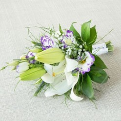 An elongated bridal-style bouquet of white lilies and purple lisianthus rests at a gentle diagonal on a pale, textured beige fabric, as if waiting to be lifted for a ceremony in Highgate. The fabric beneath has a natural, linen-like weave that sets off the flowers' colours without distraction. Several Oriental lilies open grandly at the centre, their pristine white petals curving outward like soft porcelain, dusted with delicate freckles and encircling bright yellow stamens that hold a whisper of golden pollen. Around them, sleek light green buds stretch forward, still closed, giving the arrangement a sense of unfolding time and lasting beauty. Among the lilies, clusters of lisianthus bloom in ruffled cups of white edged with vivid purple, some shading into cool lavender, others deepening toward violet, like small painterly strokes echoing evening light over Highgate Wood. Tiny sprays of white gypsophila tuck into the gaps, adding a misty, cloud-like texture that softens any hard edges and heightens the romantic tone. A full mantle of greenery supports the flowers: broad, glossy dark leaves form a strong, sculptural base, while finer wisps and spiky fronds add lift and movement, creating the impression of a carefully crafted yet organic design. At the tapered end, the stems are tightly gathered and wrapped in smooth white satin ribbon, finished with discreet pearl-like beads that suggest thoughtful craftsmanship rather than decoration for its own sake. The soft, even lighting brings out the freshness of every petal and leaf, making this bouquet feel perfect for a wedding at a local Highgate church, an engagement celebration overlooking London, or as an elegant gift delivered to a home near Parliament Hill.