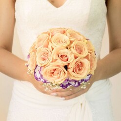 This image offers a close, intimate view of a bride's midsection, framed in soft, flattering light that feels a little like a quiet afternoon drifting over Highgate Wood. She stands poised and composed, her white or ivory wedding gown drawing the eye with its refined lace bodice and gentle V-neckline, the lace tracing delicate floral patterns that mirror the blooms she holds. The skirt falls in fluid, layered fabric, creating subtle folds that give the impression of movement even in stillness, as if she is about to step out onto a tree-lined lane near Highgate Village. Cradled carefully in both hands is a round, meticulously arranged bridal bouquet. At its centre, a generous gathering of roses in peach, apricot, and blush pink forms a lush, domed shape. The roses are tightly petalled and perfectly opened, their velvety spirals glowing with warmth and romance, like the last light over the rooftops near Pond Square. Just beneath this mass of roses, a band of ruffled purple blooms with white-edged petals-likely lisianthus-circles the arrangement, creating a vivid ring of colour that deepens the bouquet's depth and draws attention to its crafted structure. Fine, cloud-like tufts of white filler flowers, reminiscent of baby's breath, soften the boundary where bouquet meets gown, echoing the airy feel of the lace. The bride's nails are neatly manicured in a pale tone, one thumb displaying a delicate shimmer that catches the light when she shifts the bouquet. The overall impression is one of understated luxury and thoughtful design, the kind of bouquet a local Highgate florist might hand over just before a ceremony in a hidden garden or an elegant townhouse celebration, knowing it will feature in cherished photographs for decades.