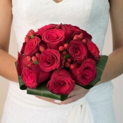 This enchanting moment, set against the leafy quietude of Highgate, encapsulates the artistry and emotion of wedding flowers at their finest. The bride's hands, elegantly manicured, support a spectacular bouquet: densely grouped, velvety red roses creating a powerful visual harmony reminiscent of Highgate's own rose-filled gardens in early summer. Accents of glossy red hypericum berries nestle between each bloom-little punctuation marks lending both festive sparkle and sophisticated contrast. Wide, structured bands of deep emerald foliage embrace the base, framing the arrangement with botanical grace and reflecting the sense of calm found beneath Highgate's canopy of mature trees. The white wedding gown-embroidered with intricate lace-gently catches the nearby sunlight, the gentle folds and textures creating interplay between fabric and floral brilliance. Each element is rendered in careful detail; the roses, full-bodied and fragrant, signal love's promise while the playful berries inject a dash of personality, as uplifting as laughter beside Pond Square. The soft, diffused light holds the arrangement in a halo, conjuring feelings of warmth, affection, and new beginnings-a fitting floral tribute for any romantic ceremony in the heart of North London.