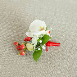 A carefully composed boutonniere lies centered on a light beige, woven fabric that resembles natural linen, its subtle texture adding a calm, understated backdrop like a table setting in a Highgate café along the Archway Road. The focal point is a flawless white Phalaenopsis orchid, its velvety petals wide and serene, cupping gently around a throat brushed with creamy yellow and soft hints of orange. This tiny burst of warm colour at the orchid's centre adds a quiet glow, drawing the eye inward. Encircling the orchid, clusters of airy white baby's breath form a delicate, cloud-like frame, each tiny blossom offering a soft contrast to the orchid's sleek, sculpted form. Just beneath, a compact cluster of round, crimson red berries glistens with a polished shine, suggesting festive joy and a hint of seasonal charm. Nestled among them, a few pale green, bead-like buds introduce a touch of early-spring freshness, as though they've just been gathered from the edges of nearby Hampstead Heath. Slender and broad green leaves weave in and out of the design, their varied shapes adding volume and a sense of natural movement without overwhelming the central bloom. All the stems are bound tightly with a rich red satin ribbon, wrapped with the kind of precision a local florist at Flowers Highgate would give to a groom's buttonhole, and tied into a neat, modest knot with short, tidy tails. The composition feels intimate and personal, perfect for weddings, proms, or formal occasions in Highgate, offering a small but meaningful accent that speaks of care, craftsmanship, and quietly confident style.