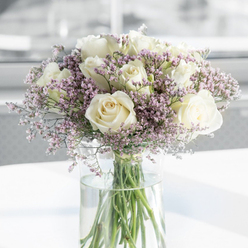 This image shows a beautifully balanced sympathy bouquet composed of lush white roses and soft lavender filler flowers, arranged in a clear glass vase on a clean, light background. Each rose is fully open, revealing a precise spiral of velvety petals that curve gently outward from the centre. The colour is a serene creamy white, with subtle ivory tones near the heart of each bloom, suggesting both purity and deep affection. Around the roses, fine, branching stems carry clusters of tiny lilac and lavender-pink blossoms-delicate, cloud-like accents that soften the overall silhouette of the bouquet. These fillers have a slightly airy, papery look, adding a gossamer layer of texture that feels particularly soothing. The green stems are trimmed and straight, all gathered within a tall, clear cylindrical vase filled with fresh water. Through the glass, the stems appear slightly magnified, their lines forming a calm, vertical pattern that contrasts with the softly rounded rose heads above. The vase sits on what appears to be a white or very pale surface, reflecting some of the gentle daylight streaming in. The background is softly blurred and free of clutter, evoking the calm, open feel of a Highgate living room with light flooding in from large windows overlooking the village or the edges of Hampstead Heath. There is a sense of quiet pause in the way the light lands on the petals and the subtle shadows cradle each bloom. The overall mood is one of tenderness and respectful remembrance-this is the kind of arrangement you might send through Flowers Highgate to console a friend, honour a loved one's memory, or bring a small, elegant comfort to a gathering after a funeral.