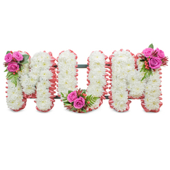 There is a tender, hands-on quality to this floral tribute that spells MUM in bold block letters, each letter densely packed with small, pristine white chrysanthemums that give a full, cloud-like texture to the surface. The letters are outlined in a soft, ruffled rose-pink ribbon that feels almost like a shawl around the white blooms, lending the piece a nurturing, intimate presence. Three small, coordinated clusters punctuate the display: one at the shoulder of the first M, one settled into the curve of the U, and one at the corner of the second M. Each cluster is composed of vivid fuchsia roses, deep green ferny foliage and tiny coral hypericum berries, their glossy skins reflecting the light and adding a delicate contrast to the matte chrysanthemum heads. A slender dark green frame is visible between the letters, discreetly supporting the standing tribute so it can be placed at a service or laid near a graveside without losing its elegant form. In a quiet studio light the flowers' textures are palpable - the mums' soft petals, the roses' velvety warmth, the berries' smooth resistance - and the combined scent is faintly floral and consoling. As a local Highgate florist who has prepared tributes for wakes at nearby chapels and the cemetery chapel, I picture this arrangement offering family members a serene focal point for remembrance, a physical expression of love that sits gently among hymns and shared memories.