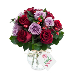 A romantic, expertly composed arrangement where crimson and burgundy roses mingle with pale lavender and mauve blooms, forming a full, rounded silhouette that suggests careful, hands-on craftsmanship. Each rose is freshly opened, its velvety petals unfurling in concentric layers, and set against deep green foliage-likely salal or Ruscus-that provides sculptural support and a cool contrast to the warm petals. Bright red-orange rose hips punctuate the bouquet, their compact, glossy spheres offering a playful, tactile counterpoint to the soft roses and adding an organic, harvest-like note. The flowers sit in a reflective mercury-glass vase with a gently rounded belly and a slightly narrowed neck; its antique silvered finish gives a soft, luminous shimmer rather than a hard shine, and a simple twine or raffia bow around the neck adds an artisanal, hand-tied feel. A small rectangular white tag hangs forward from the bow, showing a tiny stylized rose illustration and green handwritten-style script, conveying a personal greeting without overwhelming the design. Shot against a clean white backdrop, the composition reads fresh and premium-the colours saturated, edges crisp, and textures inviting touch. Imagine the faint, honeyed scent of roses with a hint of green stems and the quiet joy of receiving such a considered gift. Suited to anniversaries, expressions of gratitude, or a subtle romantic gesture, this bouquet balances classic elegance with a friendly, locally made warmth ideal for doorstep delivery across Highgate and nearby streets.