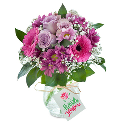 This exuberant bouquet, crafted with warmth and gentle artistry by a Highgate florist, tells a story of joy and connection through its blend of colour and form. At the centre, pale lavender roses bloom elegantly, their petals softly curled as if just caressed by the midday sun filtering through Highgate's quiet streets. Surrounding these are vivacious fuchsia Gerbera daisies, their broad, open petals and deep black centres radiating cheerfulness reminiscent of gatherings on Highgate Village green. Rich magenta chrysanthemums, some with touches of chartreuse at their centres, add complexity and texture, providing a pleasing visual rhythm. The bouquet is laced with frothy white baby's breath, bringing a sense of lightness and movement, while abundant, dark green foliage peeks through, grounding the arrangement and imbuing it with a gentle wildness akin to the wooded paths of Highgate Wood. The flowers are thoughtfully arranged in a clear vase, its cracked glass texture catching the light with subtle brilliance. A slim strand of raffia secures a simple white tag to the vase's neck-an endearing detail, suggesting thoughtful delivery and personal touch. This floral display is both uplifting and heartwarming, the perfect choice for marking special days or sending appreciation to loved ones within and around Highgate.