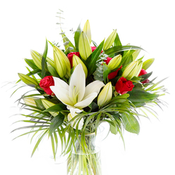 Rendered in bright, even light against a white studio field, this arrangement reads as an exercise in contrast and harmony: a single, grand creamy white oriental lily opens fully at the centre, its silky petals unfurling to display delicate pale yellow stamens and a pale green pistil, a quiet proclamation of purity. Encircling that principal bloom, many tightly held lily buds in shades of lime green and soft yellow provide rhythm and the sense of time-moments yet to bloom. Interwoven among the lilies are several small, velvety red rosebuds-each a concentrated punctuation of passionate colour-softly cupped and only beginning to reveal their inner depths. The bouquet's silhouette is shaped by luxuriant greenery: glossy broad leaves anchor the composition with deep forest tones, while lighter, arching palm fronds introduce movement as if a gentle Highgate breeze had just passed through. Sprigs of silvery eucalyptus, with their muted rounded leaves, whisper cool notes across the warmer palette and add a refined texture. The stems are visible through the clear glass vase, neatly trimmed and submerged in pristine water, reinforcing the arrangement's freshness. From a florist's viewpoint, the staging emphasizes craftsmanship-balance, repetition, and the interplay of open bloom and promise. Its scent would be a soft, intoxicating blend of sweet lily perfume and the fuller, darker note of rose. This bouquet feels appropriate for marking tender moments-anniversaries, simple celebrations, or thoughtful sympathy-and would sit gracefully on a mantel in a Highgate Village flat or as a welcoming gesture at a Hampstead gathering.
