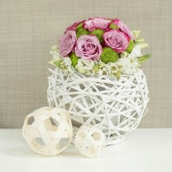 Captured in the kind of soft, even light you might find drifting through a Highgate attic studio, this image presents an artfully structured floral centerpiece designed for a wedding table or intimate dinner setting. At the base is a distinctive spherical vase fashioned from crisscrossing, twig-like strands coated in clean white, the open design reminiscent of a hand-woven basket updated with contemporary lines. From this sculptural vessel rises a dome of blooms, lush but precisely shaped. The dominant flowers are roses in shades of pale pink and muted lavender, many with slightly deeper pink edges that lend depth and a sense of ripeness. Interspersed among these are bright green button chrysanthemums, their petite, rounded forms packed with tiny petals that add both texture and a lively pop of colour, much like the fresh greens you see on a walk across Hampstead Heath. Along the lower rim of the bouquet, dainty white filler flowers create a soft fringe, easing the eye from the mass of blossoms to the basket-like vase. The ensemble rests on a smooth white tabletop that reflects the light back upward, subtly illuminating the undersides of petals and the curves of the woven strands. To the left side of the composition, two decorative orbs made from pale wicker or rattan sit casually, one slightly larger than the other, their open weaves mirroring the vase and reinforcing a theme of crafted natural materials. The background is an unfussy plane of light beige fabric, gently out of focus so the eye remains on the delicate flowers and meticulous craftsmanship. The arrangement feels calm, romantic, and perfectly at home in a Highgate dining room or local wedding venue.