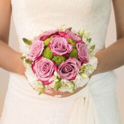 Against the detailed lacework of an ivory bridal gown, a radiant Highgate bouquet becomes a study in wedding elegance and local charm. Prominently featured are lush, full-bloomed roses in soft mauves and dusty pinks, their ruffled petals deepening to rich fuchsia along the edges-colours echoing the ancient roses found near Highgate Woods in summer. Intertwined with these, lively lime-green button chrysanthemums add a contemporary spark with their compact, dew-like form, their playful density providing an appealing counterpoint to the billowy roses. A luminous ring of small white blossoms-freesia or fine-petaled spray roses-frames the bouquet, each bud exuding a gentle, uplifting scent evocative of garden mornings. Bits of green foliage peek through, hints of woodland freshness that root the arrangement in Highgate's natural beauty. The bride's manicured hands, a gentle pink with a shimmer accent, cradle the bouquet with graceful poise, underscoring the care and intention invested by local Highgate florists. Illuminated with soft, even daylight, the entire ensemble radiates serenity, joy, and promise. Perfect for expressing the heartfelt spirit of North London weddings, this bouquet is a harmonious blend of traditional romance and playful sophistication, designed for memorable Highgate celebrations.