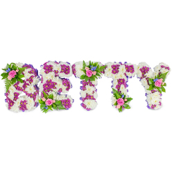 When words are hard to shape, this floral tribute speaks plainly and lovingly: the name BETTY crafted in bold capital letters from a living mosaic of flowers, set against a stark white backdrop to let every hue sing. The main fields of each letter are composed of compact, ivory-white chrysanthemum blooms that create a pillowy, tactile surface; these lend a gentle, muted scent and a sense of calm under soft chapel lighting. Inlaid and outlined within those white cushions are clusters of vivid purple-to-fuchsia chrysanthemums that form inner patterns and define the letter shapes, their deeper tones giving a subtle, dignified contrast like stitched embroidery. A narrow pale-purple ribbon trims the edges, softening the foam base and unifying the colour story. Resting on selected letters are delicate accent posies-small clusters of blush-pink roses in varying stages of bloom, accompanied by little sprays of bright blue flowers, perhaps delphiniums or cornflowers, and lacy green fern fronds. These accents punctuate the letters (notably on the B, the top of the first T, and the right arm of the Y) like gentle breaths of memory. The overall effect is serene and composed: classic sympathy whites, soulful purples, and comforting pinks balanced with fresh greenery. Thoughtfully made for memorial services in Highgate Village or beside set pieces at Highgate Cemetery, it conveys respect, remembrance, and a quietly hopeful beauty that supports families during farewell moments.