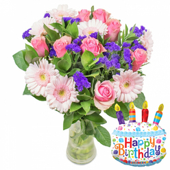 A joyful, hand-arranged bouquet captured against a crisp white studio background, the composition balancing tender romance with a lighthearted birthday flourish. The dominant voice of the arrangement is soft pink roses-petals layered like silk, shifting from a nearly translucent blush to a richer pink at the heart of each bloom-paired with generously sized pale-pink Gerbera daisies whose broad, open faces and warm yellow centers feel like a smiling welcome. Interwoven through the arrangement are tight clusters of vivid purple statice or limonium, their papery texture and compact form creating contrast and dimensionality, while small touches of creamy white filler blooms quietly brighten the bouquet's interior. Glossy, deep green foliage frames each blossom, the leaves varied in shape and sheen so the overall silhouette reads as full and natural rather than formal. All stems sit in a simple, clear glass vase with a slightly wider base, the water and stems visible and arranged to suggest freshness and care. Anchoring the scene with celebratory warmth is a cake-shaped Mylar balloon at the right-white with blue icing trim, scattered colourful sprinkles, and four striped candles in purple, red, orange and blue with tiny yellow flames-the banner across it spelling Happy Birthday in cheerful multi-coloured lettering and a small KALEIDOSCOPE mark near the bottom. As a local florist offering same-day and scheduled delivery in Highgate, near Hampstead Heath and Archway, I picture this as the perfect birthday pairing: classic blooms with a playful, modern accent for someone you love.