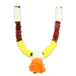 This U-shaped decorative mala, shown on a plain white field, is an exercise in rhythm, colour and cultural memory - the sort of garland you might notice in a Highgate Village window or draped above a doorway near Waterlow Park during festival season. The top of each arm is built from tight clusters of creamy-white florets that resemble small chrysanthemums; their soft, pillowy surfaces create a quiet, elegant crown where the piece is most restrained. Immediately beneath each white cap a compact strip of deep reddish-brown introduces a grounding, almost earthen counterpoint - textured, layered petals that sit like a rust-hued ribbon before the brighter bands take over. The main sweep of the garland is a vivid golden-yellow, an abundant run of marigold-like pom-poms packed so densely they offer tactile fullness and cheerful luminosity. Tiny injections of bright green mark the joins, suggesting leaves or ribbon ties and helping the eye travel through the colour sequence. At the very centre a dramatic, multi-layered orange pendant cascades down: ruffled, voluminous, and the natural focal point that draws both sight and touch. Under soft studio lighting the synthetic fibres show a slight sheen, hinting at silkiness while retaining structural resilience. As a floral professional from Flowers Highgate I describe it as ideal for puja, a temple offering, or a festive doorway - a reusable, vibrant alternative to fresh blooms for Diwali, Onam, weddings or home shrines across Highgate, Archway and neighbouring streets.