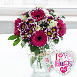 This image captures a warm, celebratory flower gift arranged as if ready to be delivered to a doorstep near Highgate Wood. In the centre, a clear glass vase sits on a clean, white tabletop, the surface reflecting soft hints of colour from above. The vase is rounded and slightly stout, filled with clear water in which the green stems are neatly aligned and trimmed, a sign of meticulous florist care. From the vase rises a full, rounded bouquet that glows with deep magenta, purple, cream and fresh green tones. Several large gerbera daisies in a rich fuchsia-magenta shade serve as the main focal points, their dark, velvety centres providing striking contrast to the bright petals. Around them, clusters of smaller multi-petalled chrysanthemums are arranged, each flower displaying deep purple petals that fade to crisp white at the tips, circling cheerful yellow centres that echo the warmth of candlelight. Creamy white roses are scattered thoughtfully throughout the design, their layered petals gently unfurling to soften the stronger colours and bring a classic, romantic note. Wispy sprigs of white baby's breath thread through the bouquet like a fine lace veil, while glossy green foliage frames the arrangement and adds depth, evoking the leafy calm of Highgate's tree-lined streets. A piece of rustic twine is tied around the neck of the vase, securing a small, rectangular white tag printed with a pink rose illustration and hand-lettered style green text, suggesting that these are handcrafted flowers. To the right of the vase, overlapping slightly, is a heart-shaped foil balloon edged in shining silver that captures glimmers of the room. Its white centre carries the words "Love n' Hugs 24-7" printed in fun, patterned letters of pink, purple and blue, some decorated with tiny hearts. The background is softly blurred and bright, likely a sunlit window, keeping the focus on this thoughtful floral and balloon set, perfect for sending love, encouragement or celebration to someone in Highgate.