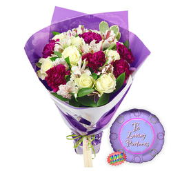Presented against a crisp white background, this romantic bouquet and balloon pairing could easily be imagined en route to a townhouse in Highgate or a cosy flat overlooking the Heath. The bouquet is generously filled, its focus resting on a cluster of creamy white roses whose petals are tightly coiled into flawless spirals, giving an impression of freshness and quiet devotion. Tucked closely among them, lush magenta and burgundy carnations bloom in dense, ruffled layers, their velvety texture adding both drama and softness. Threaded through the arrangement are stems of white alstroemeria, their trumpet-shaped flowers open to reveal intricate reddish-brown speckles and fine stripes along the inner petals, lending a painterly detail that invites a lingering gaze. Surrounding these focal blooms is an abundance of fresh green foliage, including touches of variegated leaves, which introduce subtle contrast and recall the natural greenery of Highgate's gardens and lanes. The bouquet is expertly wrapped in two layers: an inner sleeve of clean white paper provides structure and a clear line across the stems, while an outer layer of crinkled, translucent purple film envelops the flowers. This sheer cellophane rises loosely around the bouquet, catching the light and giving the blooms a soft amethyst glow. The stems at the base are neatly cinched with a slim bright green satin ribbon tied in a small bow, from which a light brown tag gently hangs, ready for a personal note. To the right of the bouquet rests a small round metallic purple balloon, its edge scalloped into a rosette shape. At its centre, a lighter purple circle contains the message "To Loving Partners" rendered in elegant pink script, while a small rainbow holographic sticker near the edge adds a flash of iridescent colour. The overall effect is of a tender, ready-made romantic gift, perfect for anniversaries, Valentine's Day, or celebrating shared milestones for couples in and around Highgate.