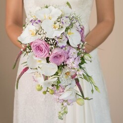There's a quiet poetry in this image-a bride, cloaked in the understated elegance of Highgate, holds a cascading bouquet that spills downward in lush, pastel abundance. The white Phalaenopsis orchids, bold yet impossibly soft, form the heart of this arrangement. Surrounding them, soft blush pink and lavender-hued roses fan out in varied stages of bloom, their layered petals catching hints of the gentle morning sun found on South Grove. Wispy threads of white baby's breath twist through the design, imparting an airy weightlessness that guides the eye along the bouquet's waterfall shape. Pops of fine magenta amaranthus tumble in textured strands, offering drama and vibrancy reminiscent of the historic gardens nearby. Subtle green foliage and unopened buds dot the ensemble, giving the bouquet both fullness and the suggestion of new beginnings. The bride's dress, with lace exquisitely detailed at the bodice, blends seamlessly into a light, muted backdrop-allowing each flower's form, colour, and texture to speak. This bouquet, crafted by Flowers Highgate, embodies the spirit of local wedding artistry: elegant, heartfelt, and touched by the natural beauty that defines this North London neighbourhood.