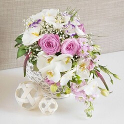A romantic floral centerpiece, ideal for a Highgate wedding or intimate dinner party, is captured in a bright, close-up view. Set in a round white wicker basket, the design showcases a lush arrangement of pink and white blooms, carefully chosen to balance softness with structure. At the heart of the piece, several large pastel pink roses are in full bloom, their spiralling petals forming classic rosettes that convey warmth and affection. Curving through and above these roses are white Phalaenopsis orchids, easily recognised by their broad, wing-like petals and glowing yellow centres, which lend the entire composition a touch of understated luxury, as if styled for a reception along Highgate High Street. Nestled among these focal flowers are airy white Lisianthus blossoms, some with delicate lavender-purple edging along their petals, introducing a gentle hint of colour variation that feels both refined and modern. Pale pink stock flowers add height and a soft vertical rhythm, their small clustered florets tumbling slightly over the rim of the basket, evoking a sense of natural movement. Throughout the arrangement, tiny clusters of white baby's breath (Gypsophila) create a fine, cloud-like texture that links each larger bloom and adds a dreamy, light effect, like morning mist over Waterlow Park. Fresh green foliage, in slender and more rounded forms, is threaded thoughtfully between the flowers, with a few leaves and buds escaping over the rim to keep the design from feeling too rigid. The white wicker basket rests on a smooth white surface, clean and uncluttered, while to one side sit two decorative balls made from intertwined natural fibres, one smaller and one larger. Their open, woven patterns echo the basket's texture and introduce a subtle rustic charm. With its neutral, softly textured background, this image beautifully conveys a mood of refinement and joy, perfectly suited to articles on wedding table arrangements, elegant celebrations, and same-day flower delivery across Highgate and nearby areas.
