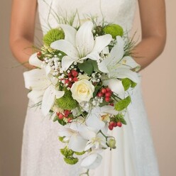 A bride stands in the heart of Highgate, her gown softly adorned with lace, cradling a breathtaking teardrop-shaped bouquet that captures both classic romance and local charm. This evocative arrangement is anchored by luminous white oriental lilies, their delicate petals curling outward and speckled faintly with reddish-brown, each bloom a study in grace and majesty. A perfectly spiraled, creamy rose sits at the centre, radiating warmth that feels at home among the blooming gardens of Highgate Wood. Gentle wisps of white baby's breath trail between the focal flowers, imparting a dreamy softness and visual airiness akin to morning mist across Waterlow Park. Verdant lime-green pom-pom mums and tufts of textured filler blooms nestle between the whites, their lively colour echoing the bright foliage lining Highgate's historic streets. Dramatic, trailing white Phalaenopsis orchids bring an effortless sense of luxury and motion to the composition, their shimmering petals falling in a graceful arc toward the bride's hands. Glossy clusters of deep red Hypericum berries are thoughtfully interwoven, punctuating the greens and whites with festive vibrancy. Framing the bouquet, slender grass-like fronds and glossy leaves offer a hint of woodland freshness, completing a design as layered and elegant as the neighbourhood itself. This striking arrangement, expertly crafted for Highgate weddings, is as memorable as a walk through its famous parks, perfectly suited for articles on seasonal bridal trends or sophisticated North London ceremonies.