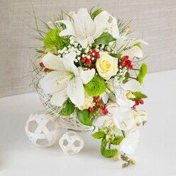 A round white wicker basket takes centre stage on a pristine white surface, overflowing with a meticulously arranged cascade of white blooms and fresh greenery, evocative of a Highgate wedding table or intimate family celebration. Rising proudly from the top of the arrangement, several large white lilies unfurl in full bloom, their trumpet-shaped petals curling gracefully backwards and revealing fine speckles and golden yellow stamens that hint at a rich, sweet fragrance. Just above the central cluster, a single cream-ivory rose sits like a quiet jewel, its layered petals forming a soft spiral that adds warmth and romance to the cooler whites. From the right-hand side, a delicate waterfall of white phalaenopsis orchids descends, each orchid bloom perfectly formed with rounded petals that overlap gently, creating a flowing teardrop effect that draws the eye downward. Around these signature flowers, the florist has woven in airy sprays of baby's breath, bright green pom-pom blooms, and textured green dianthus balls, all supported by a mix of fine, feathery leaves and broader glossy foliage that stretch outward in natural arcs. Crimson-red berries appear in small clusters, their polished surfaces catching the light and punctuating the arrangement with a festive touch, ideal for celebrations along Highgate High Street or gatherings overlooking Waterlow Park. The background features a soft, neutral beige, reminiscent of textured linen or plaster, which enhances the brightness of the whites and greens without distraction. To the left, two decorative white spheres made from interlaced strips of material rest on the table, echoing the basket's woven texture and adding a modern sculptural contrast. The entire composition feels both tranquil and celebratory, like a carefully planned centrepiece prepared by a local Highgate florist for a refined dinner party or wedding breakfast.