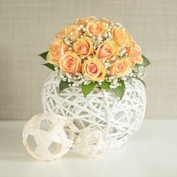 A lush, rounded bouquet of peach-toned roses forms the heart of this arrangement, each bloom nestled closely so that their velvety petals create a continuous dome of colour. The roses range from soft apricot to a richer, almost sunset orange at the core, with paler, creamy edges that catch the light and give the whole piece a gentle, luminous glow. Scattered between the roses are airy tufts of white baby's breath, their tiny blossoms creating a delicate mist that softens the shape and evokes a feeling of quiet romance, like a late afternoon walk across Hampstead Heath looking back towards Highgate. Here and there, touches of deep green foliage are tucked under the flowers, barely visible yet essential, grounding the pastel tones and adding a natural, freshly gathered feel. This floral canopy rises from a distinctive spherical vase crafted from interwoven white strips-perhaps wicker, rattan, or a similar material. The open, lattice design wraps around the stems, allowing glimpses inside while lending the piece a contemporary, sculptural quality, reminiscent of the handcrafted decor you might find in independent studios near Highgate Village. In the foreground, resting on a spotless white tabletop, sit two smaller woven spheres in a natural, beige wood tone. Their loops and crisscrossing lines echo the central vase, creating a harmonious trio that feels carefully curated. Behind everything, a light greige wall with a fine, vertical texture provides a calm, neutral backdrop, ensuring the warm colours and layered textures remain the focus. The lighting is soft and even, without harsh shadows, as if illuminated by filtered daylight in a Highgate living room. The scene conveys understated sophistication and gentle celebration, perfectly suited for an anniversary dinner, intimate wedding table, or stylish home staging across N6.