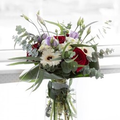 In gentle daylight reminiscent of vistas over Hampstead Heath, this bouquet unfolds in muted splendour-a tribute to the artistry found in Highgate's floral workshops. Sinuous red roses, plush and evocative, form the bouquet's heart, their deep colour softened by the luminous texture of white gerberas that mirror light drifting through ancient trees on North Road. Amidst these focal blooms, silvery eucalyptus traces arching lines, whispering a fresh, local aroma, while light mauve lisianthus-some still curled in youthful promise-emerge like delicate discoveries along Park Walk. Airy clusters of white filler flowers add volume and an almost ethereal softness, balancing the bouquet's structure with breath and space. Shining leaves cocoon the base, offering shape and a sense of continuity, while submerged stems in the glass vase reveal the life beneath. The overall impression is gentle yet assured: an arrangement equally fitting for an elegant dinner in a Highgate garden flat or as a thoughtful offering for special moments. Rose and eucalyptus blend in the air-subtle, inviting, unmistakably local-imbuing the scene with memories of springtime strolls through Highgate Woods and celebrations steeped in tradition and affection.