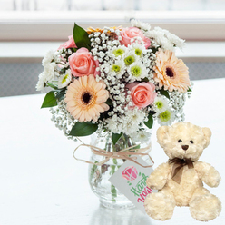On a smooth, luminous white tabletop, washed in soft daylight that suggests a north London window, a tender anniversary gift pairing has been arranged with understated elegance. The focal point is a clear glass jar vase holding a compact, rounded bouquet in soothing pastel hues. Blush pink roses, their petals unfurling like layers of silk, nestle among peach gerbera daisies whose dark brown centres create a rich, inviting depth. Surrounding these central blooms are airy clusters of baby's breath and tidy white chrysanthemums with fresh lime-green centres, giving the arrangement a light, almost cloudlike quality that calls to mind an early stroll through Highgate Wood. The stems are visible through the glass, evenly trimmed and positioned to create a graceful spiral, evidence of a skilled florist at work. Around the neck of the vase, natural twine is tied in a simple bow, and from it hangs a white rectangular tag bearing a soft pink heart and elegant green script, hinting at a handcrafted, heartfelt note. To the right, a cream-coloured teddy bear sits upright and attentive, turned slightly towards the bouquet as if sharing in the occasion. The bear's plush fur appears velvety and gently tousled, its dark button eyes and small brown nose giving it a gentle, reassuring expression. A slim brown satin ribbon is neatly tied at its neck, adding a subtle touch of formality. The background fades into a bright, out-of-focus glow, perhaps hinting at Highgate's leafy avenues or a sunlit sitting room, ensuring the viewer's eye remains on the flowers and bear. The overall mood is one of warmth, care, and quiet celebration, perfect for delivery to a partner or close friend in Highgate.