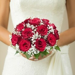 A world of tradition and emotion unfolds in this local Highgate wedding image: a bride's gentle grasp cradles a hand-tied bouquet of classic red roses, their velvety textures deepening beneath the soft light. Each rose, furled and full, nestles lovingly beside clouds of small, snowy gypsophila blossoms, whose feather-light presence lends the bouquet a sense of gentle movement. The contrast-the purity of white against crimson passion-is mirrored by fresh green leaves peeking from below, a nod to leafy Hampstead Lane and the gardens near Highgate Cemetery. Manicured hands frame the bouquet, while the wedding dress's lace-detailed fabric shimmers in ivory hues, a satin drape cascading softly to one side. The neutral, subtly blurred background lets every contour and colour sing, focusing on the quiet luxury and anticipation of the moment before the aisle. Prepared by an experienced Highgate florist, this bouquet epitomises thoughtful artistry, ideal for features on same-day bridal bouquet delivery or the enduring appeal of heritage flowers for weddings in Highgate and beyond.