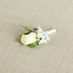 A classic wedding boutonniere is photographed on a neutral, light beige fabric that resembles finely woven linen, its subtle diagonal texture adding a calm, organic backdrop like the linens you might see set for a reception in a Highgate gastropub. Centered in the composition, yet placed diagonally across the frame, rests a single creamy white rosebud, its petals tightly wrapped and velvety, suggesting a bloom at the peak of freshness. The rose is framed by airy clouds of white baby's breath, each tiny blossom forming a soft constellation around the central flower, giving the impression of lightness and romance. Slender, pointed leaves in deep, glossy green and a few more feathery sprigs of foliage emerge from behind the rose, providing contrast and structure, reminiscent of greenery gathered from a shaded garden off Highgate West Hill. The stems of the rose and filler flowers are expertly bound together with a smooth white satin ribbon, carefully wrapped to create a clean, tailored finish. Just below the bloom sits a neat, understated bow, its loops and trailing ends lying flat, indicating the piece is ready to be pinned to a groom's or groomsman's lapel. Soft, even lighting washes over the scene, revealing the gentle curves of each petal, the delicate branching of the baby's breath, and the faint sheen of the satin, without any harsh shadows. The boutonniere exudes quiet sophistication and purity, ideal for a refined wedding ceremony or civil service in and around Highgate, and reflects the kind of meticulous, personal floristry a local wedding specialist would prepare for a meaningful, once-in-a-lifetime occasion.