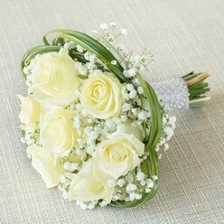 In a scene echoing Highgate's blend of stately tradition and creative flair, this beautiful bridal bouquet is displayed atop a finely woven, soft beige fabric-likely reminiscent of textiles found in quaint local boutiques. A halo of creamy white and buttery yellow roses forms the focal point, each petal softly unfurling from bud to bloom, radiating a gentle warmth that recalls tranquil mornings near Highgate Woods. The roses are generously interspersed with puffs of baby's breath, their delicate white florets lending a light-as-air quality and providing a whisper of freshness. Bright, slender bear grass weaves through the bouquet in elegant loops, framing the blooms while introducing a sense of modern movement-much like the lively spirit of Highgate's village streets. The gathered stems are enveloped in a cool silver-grey satin ribbon, meticulously adorned with rows of lustrous white pearls, offering both tactile pleasure and a refined visual touch, as if created for a bride celebrating at the heart of North London. The stems' trimmed green ends extend just beyond the wrap, adding a note of authenticity and nodding to Flowers Highgate's commitment to freshness and expertly crafted design. Perfect for a wedding, anniversary, or special milestone, this bouquet radiates purity and timeless romance, reflecting both the elegance and warmth found throughout the Highgate community.