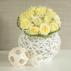 Set against a soft, neutral backdrop, this image focuses on a beautifully crafted wedding-style centerpiece that could easily grace a Highgate reception table. At the core is a full, dome-shaped bouquet of pale yellow roses, the blooms placed so close together that they create a smooth, almost sculpted surface of petals. Each rose is fully open, revealing ruffled layers that seem to glow softly, like early sunshine over the rooftops near Highgate Village. Threaded through this sea of yellow are sprigs of white baby's breath, their countless small flowers forming airy tendrils that lighten the design and introduce a whimsical, cloud-like texture. Around the base of the bouquet, a slender ring of vivid green foliage has been laid with precision, providing a crisp band of colour that separates the floral mound from the container and enhances the sense of structure. The vase is a striking white woven form, rounded and slightly cinched at the middle, reminiscent of a modern basket made from interlocking strands. Its open weave allows glimpses of shadow and negative space, which play across the smooth white tabletop and give the arrangement a feeling of lightness and ease. Nearby sit two decorative spheres fashioned from pale, natural-looking strips arranged into open, looping frameworks, echoing the same woven motif and lending an extra layer of textural interest. The softly blurred background in warm beige creates a tranquil, gallery-like setting where the centerpiece can shine. The overall impression is of calm sophistication, ideal for a wedding near Highgate Wood, an anniversary dinner at home, or any event where subtle luxury and thoughtful design are desired.