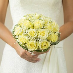 Evoking the classic gentility of Highgate, this bridal bouquet is a celebration of light, texture, and refined local craftsmanship. The image offers an intimate view of a bride's gently clasped hands, her nails painted in a soft nude, accentuating the round bouquet she holds with care. Each yellow rose within the arrangement is meticulously selected for its creamy hue and folded perfection, the petals swirling like silk ribbons beneath the high canopy of a white wedding gown. Wispy clouds of baby's breath drift between the roses, lending the bouquet a delicately ethereal quality reminiscent of early morning mists rolling over Highgate Wood. Bound together by thin bands of dark, lustrous foliage, the bouquet gains a sculptural form and quiet confidence. The subtle texture of the bridal gown and the gently blurred background ensure the flowers remain center stage, inviting every viewer to appreciate the artistry and anticipation layered in each detail. This floral arrangement captures Highgate's unique sense of place-elegant, historic, and full of promise-making it perfect for a bride who appreciates both tradition and contemporary floral design.
