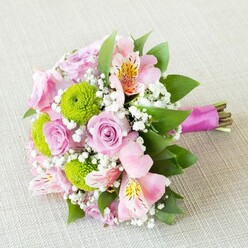 Cradled lovingly on a softly textured cream fabric, this bouquet channels the serene elegance of Highgate's leafy avenues. The pastel pink roses, each blossoming in gentle stages from tight buds to fully opened blooms, create a sense of unfolding romance and promise. Scattered among these are playful lime-green button chrysanthemums, their compact spherical heads providing exuberant pops of colour and a tactile contrast reminiscent of Highgate Wood in spring. A touch of the exotic arrives in the form of pink alstroemeria blooms, their slender petals dancing with strokes of yellow and dark burgundy, weaving delicate patterns across the bouquet. Wisps of airy white baby's breath settle between the blossoms, giving the arrangement a sense of lightness and breathability, like a gentle morning in Waterlow Park. Glossy green foliage peeks from beneath and between the flowers, grounding the bouquet in the local landscape and adding depth. Secured at one end, a ribbon of fuchsia satin is tied snugly around the stems-its vivid hue boldly complementing both flowers and leaves. The entire creation is lit by gentle, natural light that coaxes out the soft variations in texture and colour, making each element sing. Designed for moments of celebration-whether a wedding or a heartfelt gift for a loved one in Highgate-this bouquet combines artistry, freshness, and the unmistakable warmth of a community florist.