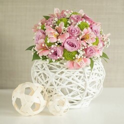In this carefully composed image, a voluminous floral centerpiece stands confidently on a spotless white tabletop, bathed in soft, even studio light that mimics a gentle Highgate morning. At the heart of the scene is a distinctive white vase, created from slim, interlaced strips that form an open, basket-like lattice. Its rounded, weighty shape suggests stability, the kind of piece you might find gracing a well-loved home on a quiet lane off Highgate High Street. Spilling generously from the vase is a rounded bouquet of pastel pink and lavender roses, each bloom fully unfurled, revealing velvety petals arranged in intricate spirals. Nestled among them are clusters of pink alstroemeria, recognisable by their slender petals brushed with fine stripes and tiny freckles of deeper colour, bringing a slightly wild, garden-fresh note to the composition. Lively lime green button chrysanthemums dot the arrangement like beads of energy, their tight, pom-pom heads offering crisp contrast against the soft blush tones. Wisps of delicate white baby's breath weave through the design, cloud-like and weightless, creating a sense of breath and movement, as if a light breeze from the slopes of Parliament Hill had passed through. Rich green foliage peeks out between the flowers, framing the pastel palette and grounding the composition. To the left of the vase, two decorative woven spheres in natural beige tones rest on the table, their airy, rattan-style construction mirroring the texture of the vase and completing the vignette with a contemporary, handcrafted feel. The overall effect is of quiet celebration and gentle sophistication, ideal as an elegant centerpiece for a Highgate dinner party or a same-day floral delivery to mark a tender anniversary at home.