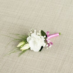 A single white lisianthus boutonnière lies on a warm, sand-coloured fabric that resembles natural linen, its woven texture adding a rustic yet refined backdrop often favoured for weddings in Highgate's elegant townhouses and small chapels. The central bloom is fully opened, its soft, ruffled petals layered in concentric circles that create a gentle, cupped shape, like a tiny piece of sculpted porcelain. Just to one side, two elongated lisianthus buds, creamy with a hint of pale green, rest together as if in conversation, echoing the theme of partnership and new beginnings. Fine sprays of white baby's breath thread through the design, their tiny blossoms catching the light like scattered pearls, adding airiness and volume without overshadowing the focal flower. Behind the blooms, a single deeper green leaf anchors the composition, while several slender, grass-like blades arc gracefully towards the lower left corner of the image, introducing a sense of movement similar to a breeze through the trees on Hampstead Heath. At the base, the stems are wrapped in a vivid fuchsia satin ribbon, tied into a neat bow that delivers a bold splash of colour against the otherwise soft palette. The sheen of the ribbon suggests careful, expert hands at work, like those at Flowers Highgate preparing a groom's buttonhole for same-day delivery before an afternoon ceremony. Soft, even lighting highlights the freshness of the petals and the crisp contrast between white, green, and pink, conveying an overall feeling of refined joy and quiet celebration.