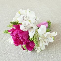 A small but luxurious floral arrangement lies gently on a pale beige, linen-like cloth, as if set out on a dressing table in a Highgate townhouse before a wedding. The eye goes first to the lush central bloom: a fully opened peony in a deep fuchsia-magenta shade, its many ruffled petals pressed tightly together, creating a soft, romantic fullness. Surrounding this main flower are several white alstroemeria, or Peruvian lilies, each with six slender petals that curve slightly back, revealing throats delicately patterned with fine maroon streaks and speckles. These markings add intricate detail, like hand-drawn lines on porcelain. Threaded between the larger flowers are tapered green buds and spiky florets, perhaps from gladiolus or similar filler stems, bringing a fresh, vertical texture and a vivid pop of spring green. The stems are neatly gathered and wrapped in a smooth, bright pink satin ribbon that mirrors the intensity of the peony and adds a contemporary, polished touch. The neutral, softly textured fabric beneath provides a calm backdrop, allowing the rich colours to glow and the shapes to stand out clearly. Gentle, diffuse light highlights the satin sheen of the ribbon, the subtle shadows along each petal edge, and the natural variation in the greens. The arrangement feels like a bespoke boutonnière or corsage crafted for a celebration near Highgate Wood, ideal for bridal parties, prom nights, or intimate celebrations at local restaurants, its compact size and bold palette expressing effortless joy and modern romance in one carefully tied cluster of blooms.