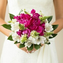 In this detailed bridal close-up, the camera lingers on a hand-tied wedding bouquet held carefully against the bodice of a white lace gown, capturing a moment of stillness before the walk down the aisle. The bouquet's core is a lush cluster of magenta and fuchsia peonies, each bloom sumptuous and full, with tightly layered, ruffled petals that give a sense of depth and luxury. Mingling between them are crisp white alstroemeria, their petals flared open to reveal intricate freckles of dark maroon along the throats, lending both contrast and a touch of exotic charm. Slim, spiky green buds and unusual textural elements, perhaps Ornithogalum or similar filler flowers, peek through the composition, their fresh green tones and upright forms adding energy and lightness. A collar of broad, deep green leaves encircles the bouquet, providing structure and framing the colour palette so that the peonies' intense pinks seem even more striking. The bride's hands, nails softly polished in a pale blush, wrap around the stems, and the soft folds of her gown create a gentle, neutral backdrop. The overall lighting feels natural and airy, reminiscent of a ceremony space near Highgate Wood or a bright room overlooking the village streets. The image conveys both refinement and exuberance, suggesting a bride who loves classic romance but isn't afraid of vibrant colour. It's the kind of bouquet a local Highgate florist might recommend for a spring or early summer celebration, when peonies are at their freshest and their fragrance can subtly perfume each step up the aisle.