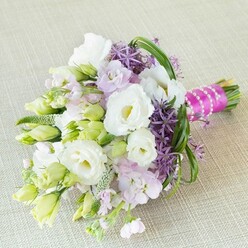 Washed in soft, natural light and resting gently on a plush cream background, this hand-tied bouquet bursts with optimism and charm-qualities cherished by the Highgate community. Full white Lisianthus blossoms, opening to reveal sunlit, ruffled petals, intertwine with tightly closed buds that hint at promise and anticipation. Airy pastel stocks in lavender-pink weave gentle fragrance and dreamy color through the arrangement, capturing a hint of garden pathways near Highgate Wood. Offsetting these softer blooms, clusters of vibrant deep purple allium command attention with their unmistakable star-shaped florets. Throughout, sprigs of lime-green Lisianthus buds and delicate, textural greens evoke a sense of natural growth and renewal, reminiscent of Waterlow Park's first flushes of spring. Slender arcs of glossy bear grass outline the bouquet, imparting graceful structural movement. At the stems, a striking hot pink satin ribbon, finished with an elegant strand of gleaming pearls, adds celebratory flair and personality to the design-a touch that would be at home at a wedding in one of Highgate's historic venues. Radiating with the quiet sophistication and heartfelt unity that define local gatherings, this bouquet, thoughtfully crafted by Flowers Highgate, serves as a symbol of artistry, romance, and joyous milestones.