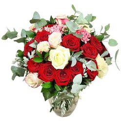 A lavish rose bouquet fills a clear glass vase, presented against an immaculate white background so that every hue, petal, and leaf feels intensified, much like an elegant arrangement arriving at a Highgate home near Dartmouth Park Hill. The bouquet overflows with approximately two dozen or more roses, forming a generous, dome-shaped display. Deep red and bright scarlet roses dominate the centre, some tightly furled in bud and others fully unfurled so their velvety petals billow outward in layers, suggesting deep love and celebration. Interspersed among these passionate reds are creamy white, ivory, and softly blush-tipped white roses, which cool and soften the palette, adding an air of purity and gentle warmth. Dotted throughout are small clusters of vivid pink filler blooms, reminiscent of mini carnations or similar flowers, giving the arrangement extra texture and subtle pops of lighter colour. Wisps and arcs of blue-green eucalyptus weave in and around the roses, their rounded, silvery leaves lending a fresh, contemporary feel and suggesting a clean, aromatic scent drifting through a Highgate sitting room. Dark green foliage and occasional silvery-green leaves frame the blooms and fill in the spaces, creating a balanced, rounded shape that appears lush from every angle. The clear glass vase below has a slightly bulbous base tapering into a narrower neck, allowing the stems to be seen through the water, adding an honest, garden-picked charm to the otherwise luxurious display. With the stark white background stripping away distractions, the bouquet becomes a striking symbol of romance and elegance, ideal for anniversaries, proposals, or any heartfelt occasion celebrated in and around Highgate.