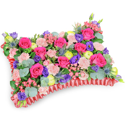 Taken from a slightly raised, angled viewpoint on a pure white surface, this rectangular pillow of blooms feels like a carefully composed keepsake from a Highgate florist's hands. At its heart are numerous hot pink roses-sumptuous and velvety-ringed by soft, shell-pink spray carnations that create a plush, inviting topography. Bright lemon-yellow ranunculus scatter like small suns, while deep purple lisianthus (eustoma) adds a satin-dark contrast that reads as depth and dignity. The silver-blue spikes of eryngium introduce a wild, architectural whisper among the softer blossoms, and tiny pale-pink filler flowers knit the composition together with delicate, lace-like punctuation. Rounded eucalyptus leaves in a cool silvery-green and other lush foliage lie beneath and between the blooms, giving the cushion substance, natural movement, and a subtle scent that lifts in the air. Around the base, a pleated deep-red satin ribbon forms a ruffled frame-both formal and comforting-defining the pillow's shape with tailored finesse. As a memorial or sympathy tribute, this arrangement leans toward celebration of memory; the saturated fuchsia and lemon tones speak of joy remembered as much as loss felt. Crafted with the kind of attention we use at Flowers Highgate for local deliveries across Highgate Village and near Waterlow Park, the piece balances technical structure with emotional warmth, offering a fragrant, visual embrace that would sit peacefully on a chapel table or beside a family photograph.