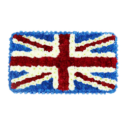 This image shows a carefully composed funeral tribute in the shape of a perfect rectangle, depicting the United Kingdom's Union Jack entirely through flowers rather than fabric. Set against a clean white backdrop, the floral flag stands out with striking clarity, every line and colour thoughtfully placed. The background is made from a dense mass of deep blue blossoms, likely hydrangeas or similar compact flowers, trimmed to form a smooth, uniform field that fills each corner and creates a neat frame. On top of this blue base sits the commanding red St George's Cross, its vertical and horizontal stripes crafted from tightly packed red chrysanthemums or button carnations that form a bold, unbroken block of colour. From corner to corner, the narrower red diagonals of St Patrick's Cross slice through the design, aligned with such precision that each edge remains crisp. Bright white blooms create the diagonal St Andrew's Cross and trace slim borders around all the red elements, providing the characteristic fimbriation that defines the Union Jack's structure. The close clustering of petals lends the tribute a plush, almost cushion-like texture, as if the flag might yield softly at a touch. The overall effect is solemn yet proud, conveying deep respect and shared heritage. In Highgate, an arrangement like this might be chosen for a service connected to military history or public service, perhaps for a ceremony close to Highgate Cemetery or a memorial gathering along Highgate Hill, where friends and family wish to honour a life intertwined with British traditions and values.
