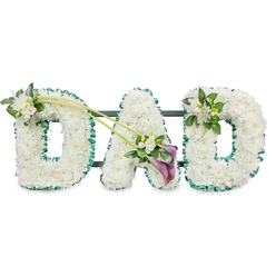 A refined "DAD" funeral tribute stands out against a plain white background, its simple setting allowing every floral detail to be clearly appreciated. The three letters are large and upright, densely filled from edge to edge with pristine white chrysanthemums or similar small blooms. The petals overlap to create a soft, cushioned surface, like freshly fallen snow, suggesting purity and the tenderness of cherished memories. A ruffled band of deep green satin ribbon outlines each letter, providing a vivid contrast that echoes the leafy avenues near Highgate Cemetery and Waterlow Park. The structure behind the flowers is subtly visible in places, with slim dark bars connecting the letters and indicating a strong frame that lets the tribute stand securely during a funeral or memorial service. On the upper left curve of the first "D," a delicate cluster of white freesia and polished green foliage is placed thoughtfully, as if gently resting on the chrysanthemum bed. Two fine green stems arc from this cluster across towards the central "A," where they open into an expressive spray. Within this focal area, two rich, deep purple calla lilies curl gracefully among white blossoms and leafy greens, their trumpet-shaped blooms adding both sculptural interest and a deeper emotional tone. The final "D" on the right bears a smaller matching arrangement of freesia and foliage at its top right edge, mirroring the first letter without overpowering the design. The result is a personalised tribute that feels both intimate and dignified, suitable for honouring a father at services across Highgate and the surrounding N6 community.