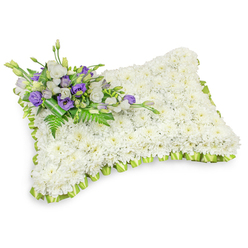 This floral pillow reads as a peaceful, restorative token: a rectangular cushion shape formed entirely from a dense carpet of immaculate white chrysanthemum heads, each blossom packed closely to create a uniform, velvety texture that suggests softness and rest. The piece is edged in a pale lime-green ribbon, expertly pleated into a continuous scalloped trim that traces the pillow's rounded corners and gives the design a gentle, uplifting outline. In the upper-left quadrant a composed cluster of accent blooms rests like a quiet thought - white lisianthus with soft, ruffled layers beside deep purple lisianthus, several kept in bud to add a sense of reserve and unfolding memory. Sprays of feathery fern fronds and deep green leaves are woven through this accent, and a darker green leaf wrap at the base holds the stems neatly, presenting an elegant, finished hand. Photographed against a stark white background with a soft shadow beneath, the arrangement reads three-dimensionally, as if ready to be carried to a graveside or placed by a memorial card. The scent is implied rather than overwhelming, the texture tactile - the chrysanthemums' cool, papery petals, the lisianthus' silky cups, the ribbon's satin sheen - all contributing to an atmosphere of consolation. For families in Highgate and the surrounding streets such as North Hill and Swains Lane, this pillow offers a comforting, traditional expression of sympathy and remembrance, made with careful craftsmanship and an eye for quiet, respectful detail.