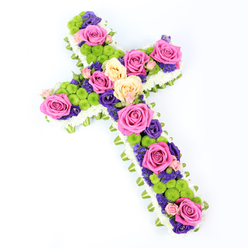 This photograph shows an elegant, cross-shaped floral tribute, laid out on a pure white background that allows each colour and texture to stand out with clarity. Viewed from a modest overhead angle, the cross appears slightly tilted from upper left to lower right, giving the arrangement a dynamic yet gentle presence, as though ready to be carried into a Highgate chapel or service near Archway Road. The outer edge of the cross is meticulously formed from densely packed white spray chrysanthemums, their small, rounded faces creating a soft, plush border that speaks of peace and purity. Just below this cushion of white, a narrow band of folded pale green ribbon or foliage wraps the perimeter, adding a subtle halo of fresh colour, reminiscent of the foliage lining the paths through Waterlow Park. Inside this defined edge, the central area of the cross is rich with colour. Large, open roses in vivid fuchsia pink and soft creamy yellow are strategically placed to draw the eye along the vertical and horizontal arms, their velvety petals unfurling in full bloom. Between the roses, bright lime-green button chrysanthemums have been tucked in clusters, their compact, spherical heads offering playful texture and a burst of contemporary freshness. Weaving through these elements are smaller flowers in deep purple and velvety violet - likely lisianthus or statice - which lend depth, contrast, and a sense of quiet reverence that feels appropriate for a funeral or memorial. The lighting is bright and even, with minimal shadow, ensuring every petal, ribbon fold, and green accent is beautifully defined. Crafted with the sensitive touch of an experienced local florist serving Highgate, this floral cross expresses both gentle sympathy and a hopeful celebration of life and faith.